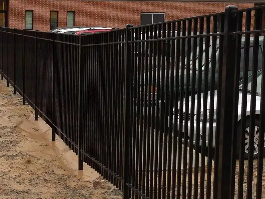 A black metal fence with vertical bars stands on muddy ground, separating a parking lot with several cars from a brick building in the background.