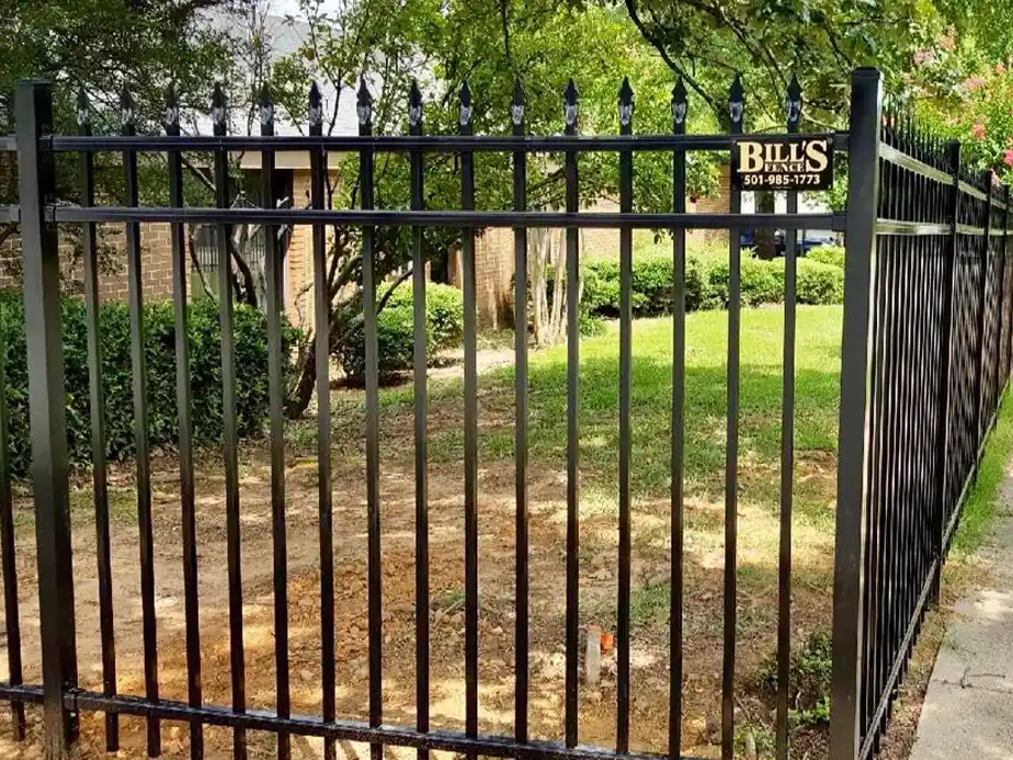 A black metal fence with pointed tops surrounds a yard with grass, trees, and bushes. A sign on the fence reads “Bill’s” with a phone number. The background shows part of a brick house.