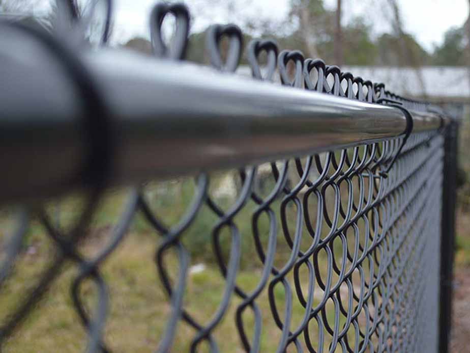 Close-up view of a black chain-link fence, with the camera focused on the top rail and wire links. The background is blurred, showing some greenery and indistinct outdoor features.