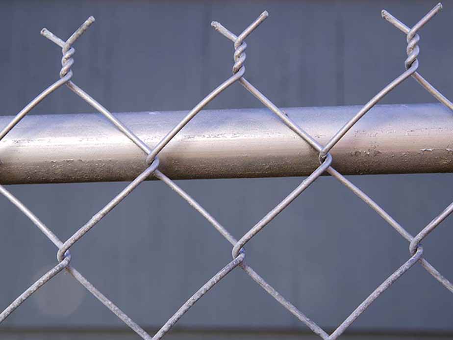 Close-up of a silver chain-link fence with twisted wire tops, positioned in front of a horizontal metal pipe and a blurred gray background.