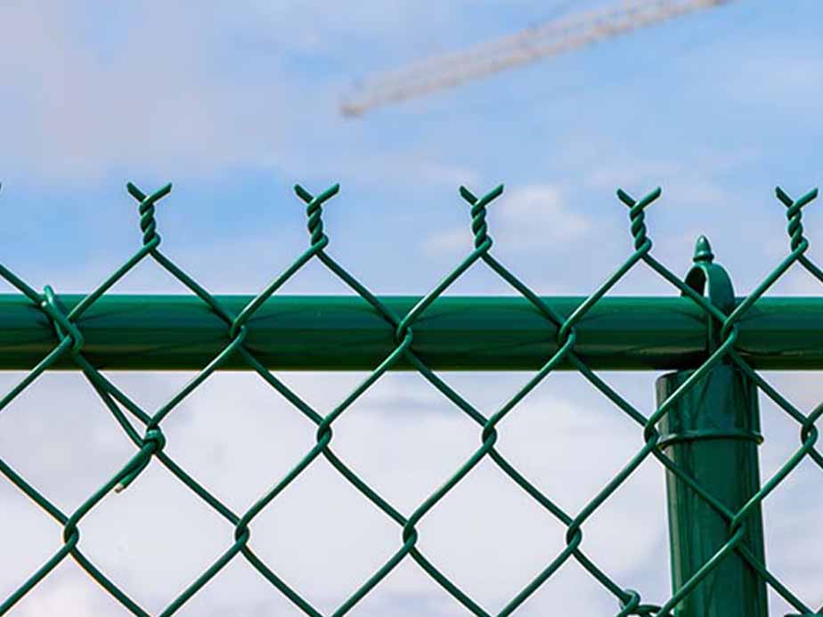 Close-up of a green chain-link fence with barbed wire on top against a blue sky with scattered clouds. A blurred structure, possibly a crane, is visible in the background.