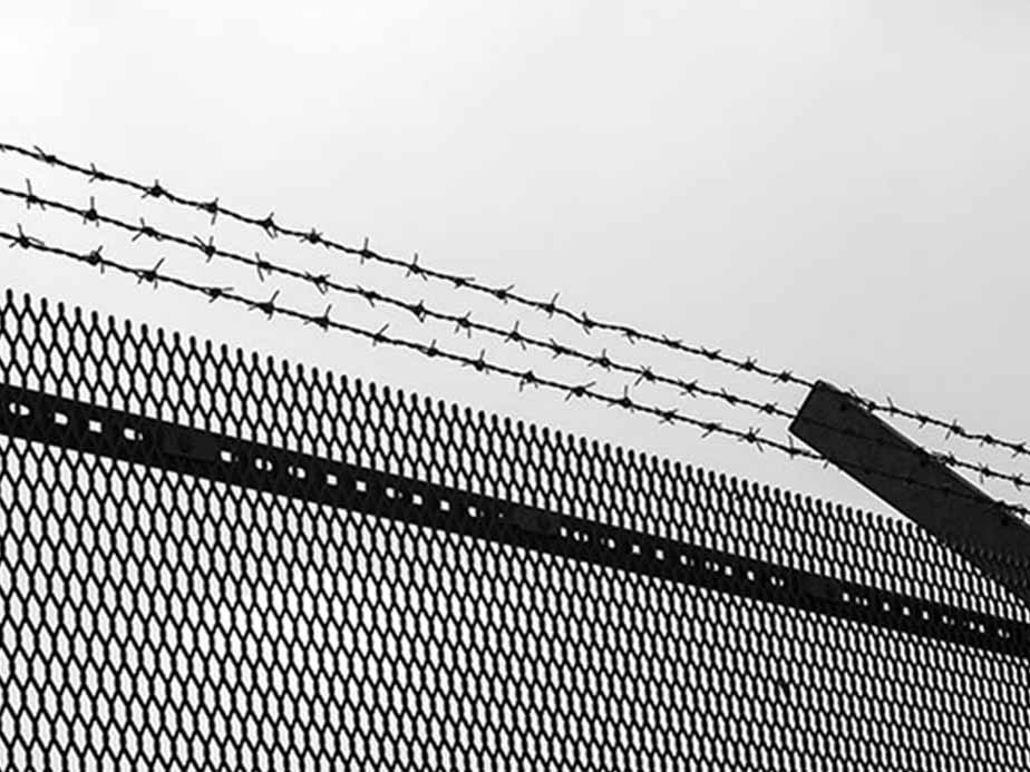 A black and white photo of a metal fence topped with angled strands of barbed wire, set against a plain, overcast sky. The image conveys a sense of security or restriction.
