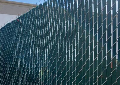 A green privacy screen is attached to a chain-link fence, creating a solid barrier. The top of the fence has angled metal posts and barbed wire, and the sky is visible in the background.