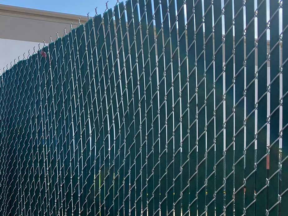 A green privacy screen is attached to a chain-link fence, creating a solid barrier. The top of the fence has angled metal posts and barbed wire, and the sky is visible in the background.