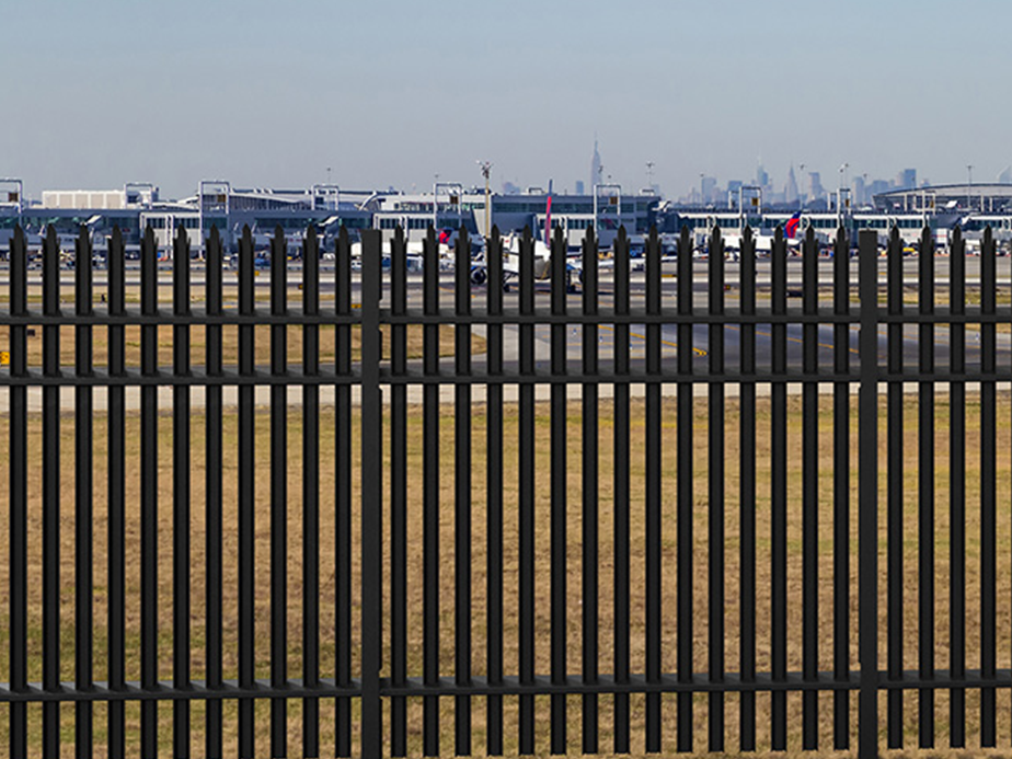 A tall black security fence in the foreground separates a grassy area from an airport runway with airplanes and terminal buildings visible in the background. The city skyline is faintly visible in the distance under a clear sky.