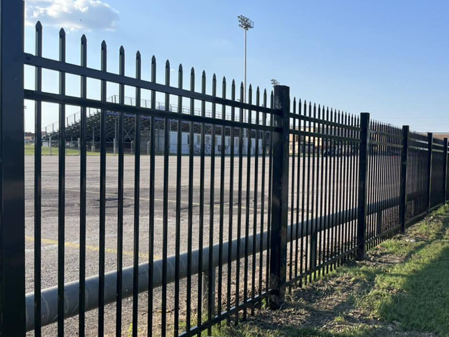 A black metal fence with pointed tops runs alongside a grassy area, enclosing an empty parking lot. In the background, there are stadium bleachers and tall light poles under a clear blue sky.