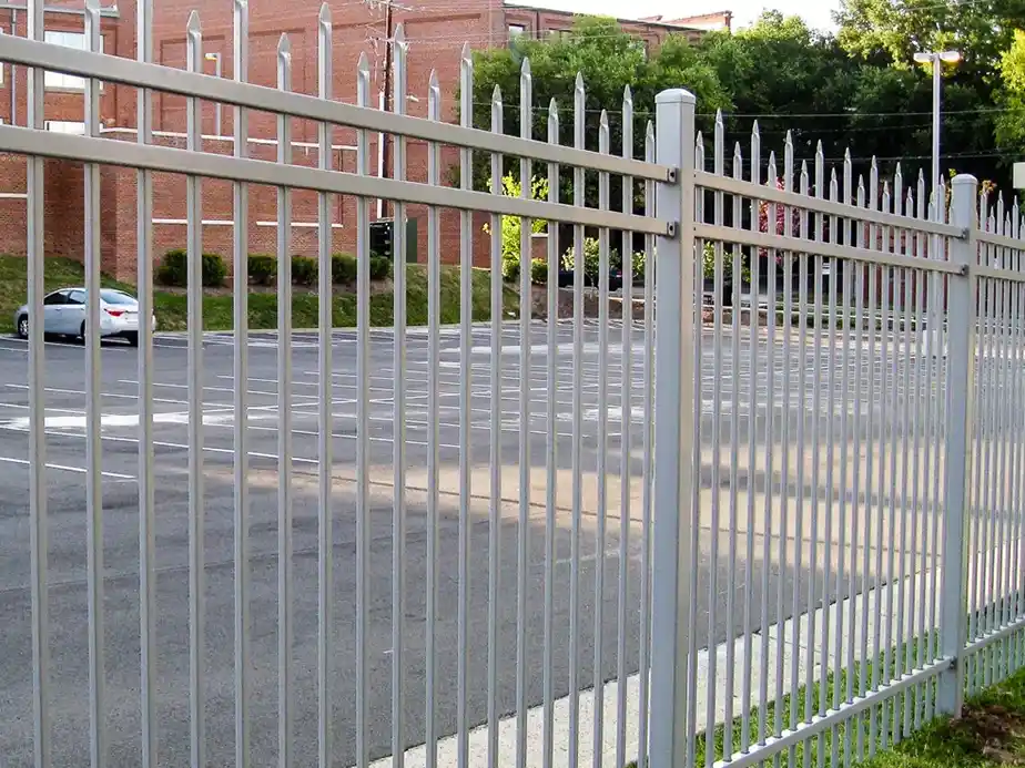 A metal security fence with pointed tops surrounds an empty parking lot. In the background, a red brick building, trees, and a parked car are visible.