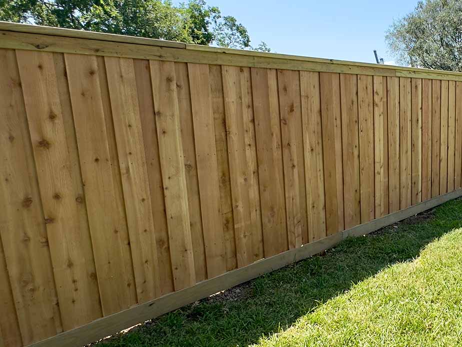 A wooden privacy fence stands on a grassy lawn, with trees and a clear blue sky visible in the background.