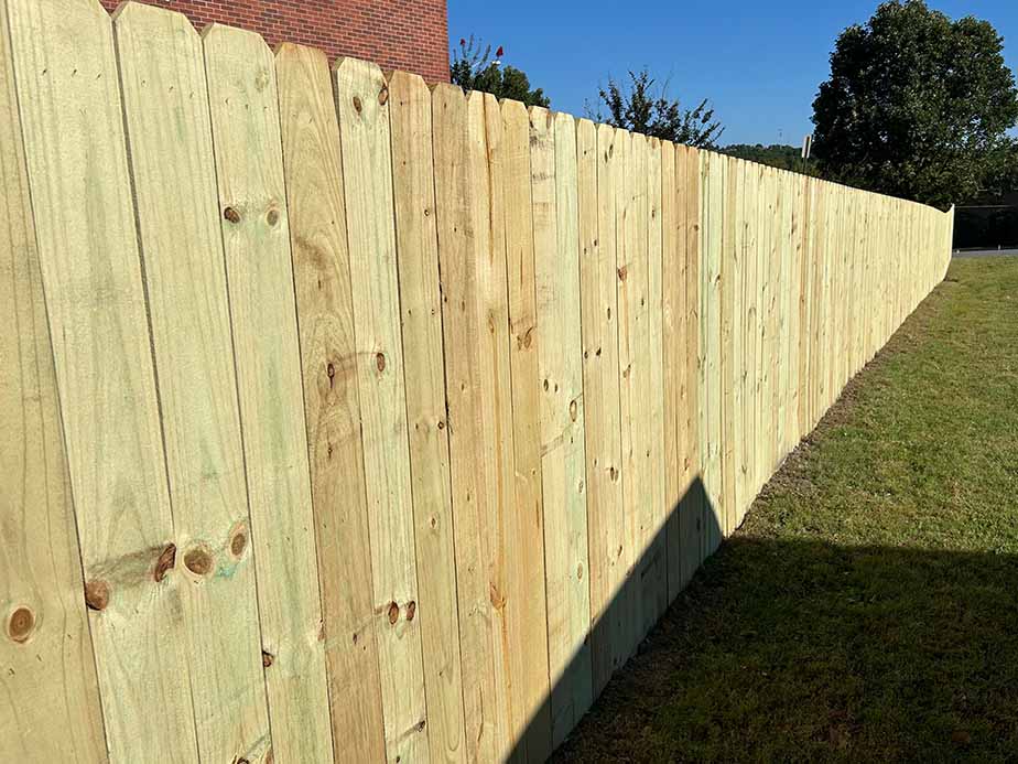 A long, wooden privacy fence runs alongside a grassy yard, casting a shadow on the grass. Trees and part of a brick building are visible in the background under a clear sky.