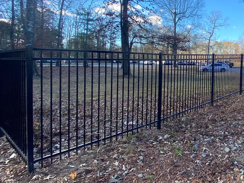 A black metal fence runs across a leaf-covered area with trees and parked cars visible in the background under a blue sky. The fence ends abruptly, not enclosing anything.