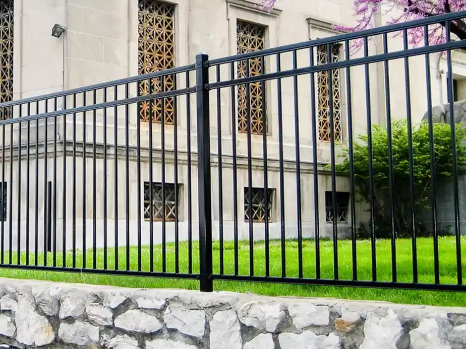 A black metal fence stands in front of a stone wall, with a green lawn and a beige building featuring ornate window grills in the background.