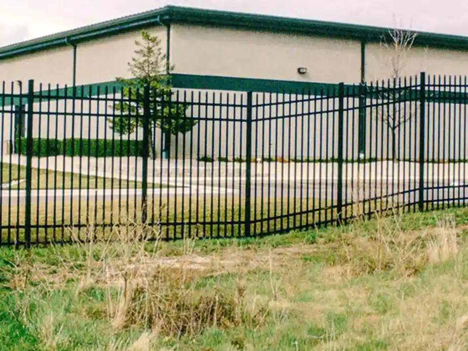 A black metal fence with a bent section stands in front of a beige industrial building with a green roof. There is grass and some dry weeds in the foreground.