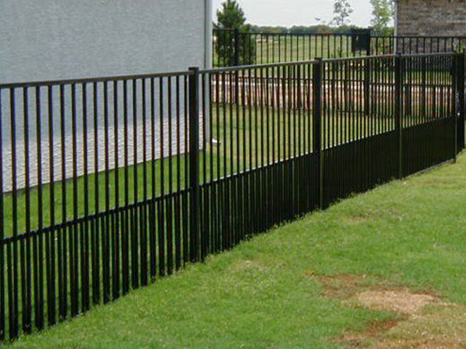 A black metal fence with vertical bars runs along a green lawn beside a gray building, with trees and open field visible in the background.