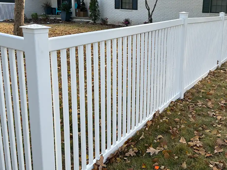A white vertical slat fence runs along the edge of a yard with green grass and fallen autumn leaves, in front of a white brick house with shrubbery and trees.