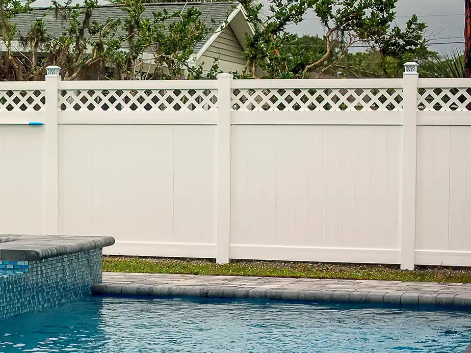 A white vinyl privacy fence with a lattice top borders a backyard swimming pool, with green grass and trees visible behind the fence.