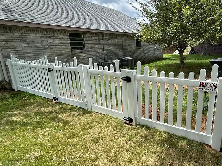 A white picket fence with a gate stands between a brick house and a grassy yard, with a tree nearby and an HVAC unit next to the house. A sign on the fence displays a business name and phone number.