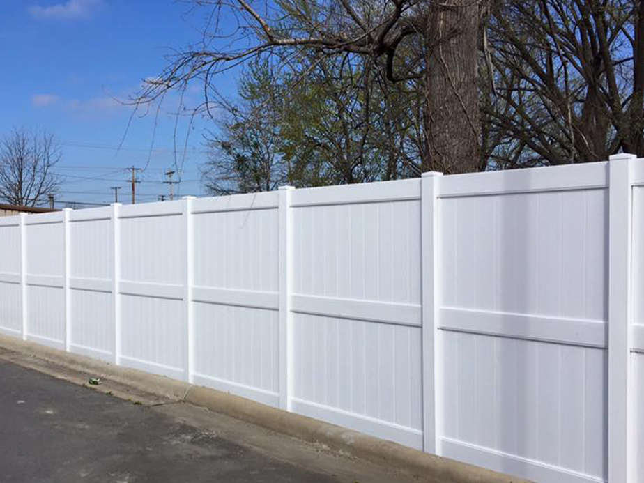A tall, white vinyl privacy fence runs along a paved road, with bare and leafy tree branches visible behind it under a blue sky.