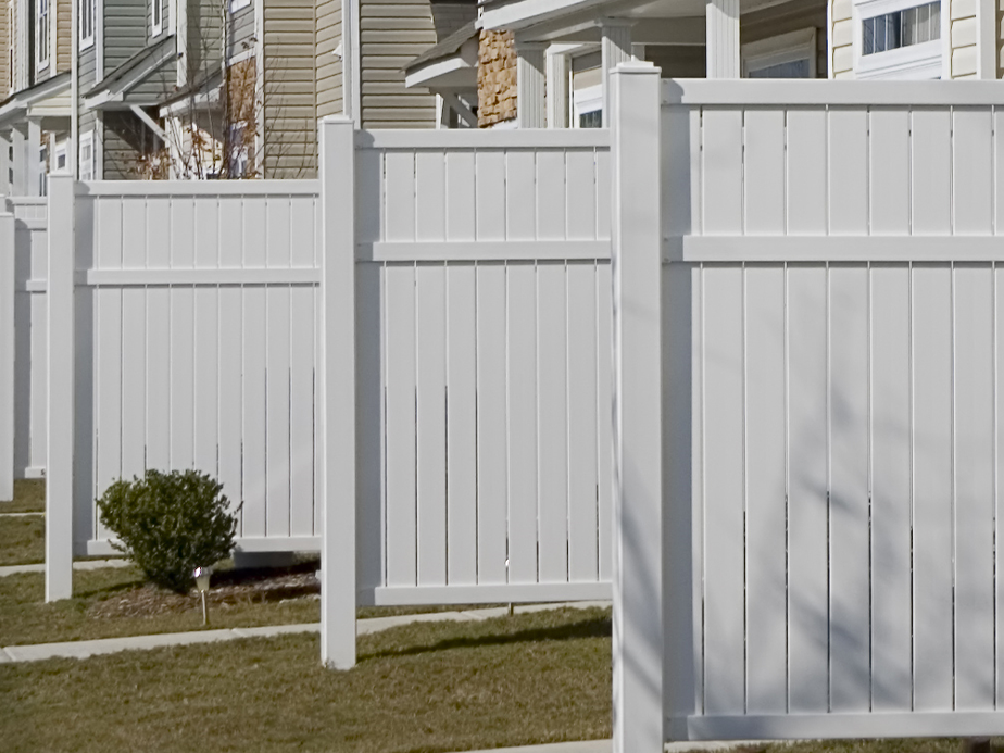 A row of white vinyl privacy fences separates the yards of modern suburban houses, with green grass and small shrubs visible along the fences.