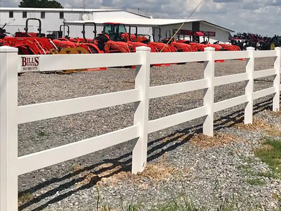 A white vinyl ranch-style fence runs along a gravel lot with several orange tractors parked in rows near a white building under a partly cloudy sky.