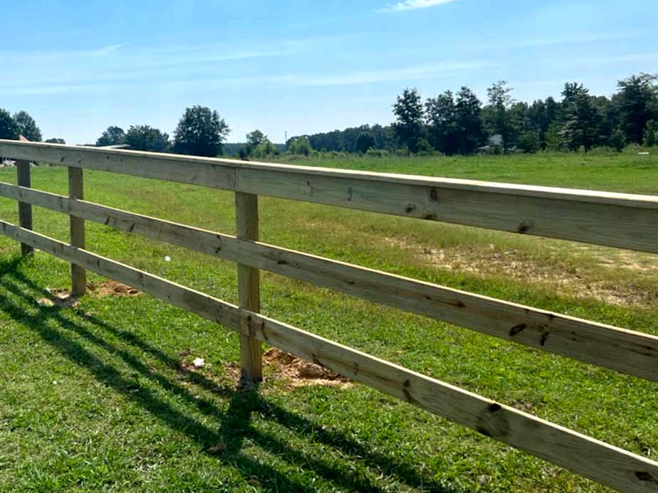 A wooden fence runs diagonally across a grassy field under a clear blue sky, with trees in the background and shadows of the fence cast on the grass.