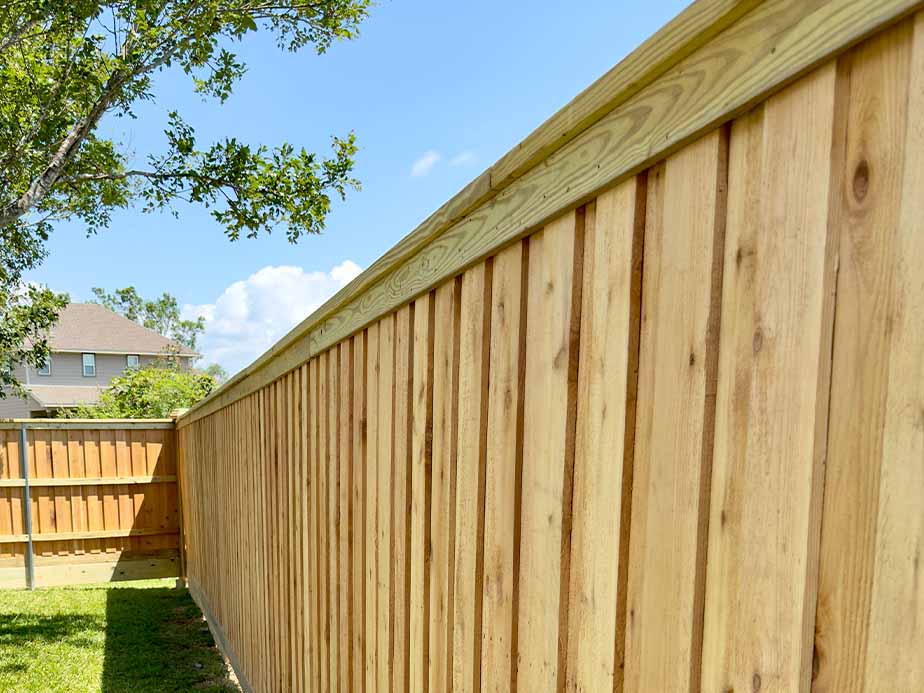 A tall wooden privacy fence runs along a grassy backyard, with a tree and a house visible in the background under a bright blue sky.