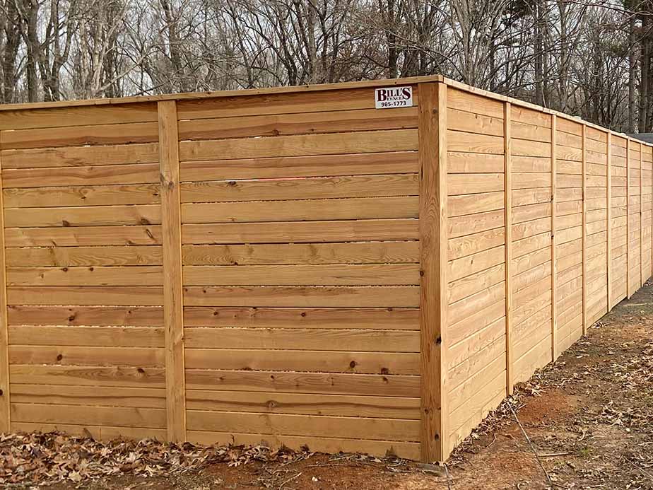 A tall wooden privacy fence with horizontal planks surrounds an area outdoors. The ground is covered with dry leaves and dirt, and bare trees are visible in the background. A small sign is attached to the top of the fence.