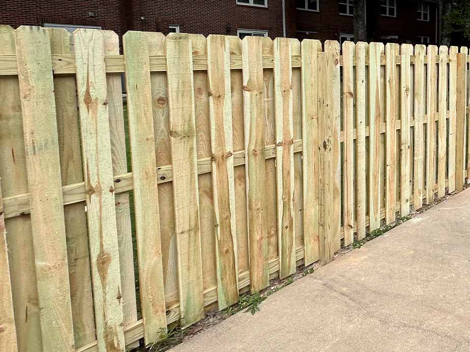 A wooden picket fence with vertical slats stands next to a concrete sidewalk. There are grassy patches at the fences base and a brick building is visible in the background.