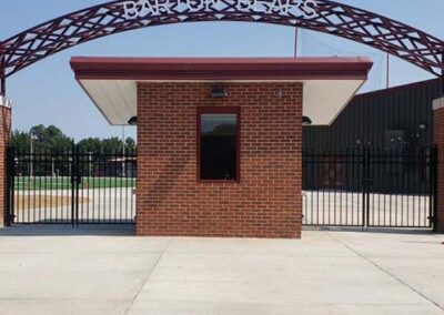 A brick ticket booth stands in front of a black metal gate, with a curved maroon arch above it that reads “Barton Bears.” Trees and sports fields are visible in the background.