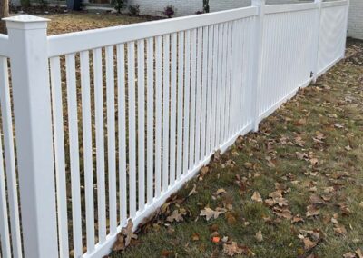 A long white vinyl picket fence runs alongside a yard with patchy grass and scattered brown autumn leaves. Trees, bushes, and part of a white brick house are visible in the background.