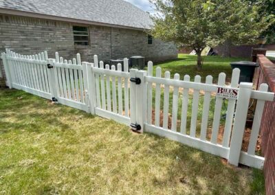 A white picket fence with a gate stands in a grassy yard next to a brick house and a tree. There is a sign on the fence that says Bills with a phone number.
