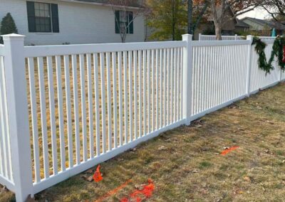 A white vinyl picket fence runs along a grassy yard in front of a house. The fence has vertical slats and a few holiday wreaths. Orange spray paint and small flags mark the ground near the fence line.