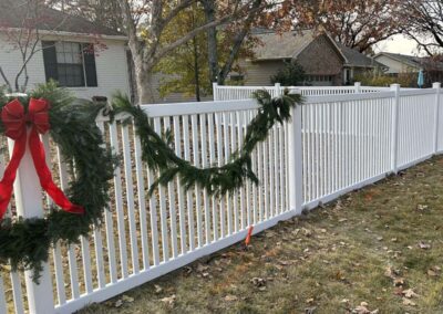A white picket fence decorated with a large green wreath adorned with a red bow and a garland of greenery. Houses and trees with autumn leaves are visible in the background.