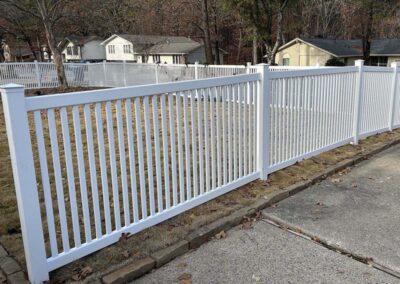 A white vinyl picket fence runs alongside a sidewalk and encloses a grassy yard, with houses and trees visible in the background.
