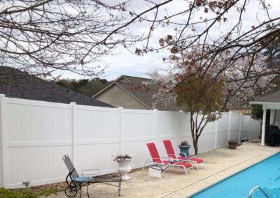 A backyard scene with a swimming pool, three red lounge chairs, and two white planters beside a tall white privacy fence. Leafless tree branches hang overhead, and rooftops are visible in the background.