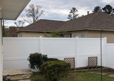 A backyard with a white vinyl privacy fence, green bushes, and small lattice panels along the fence. A neighboring house with a brown roof is visible behind the fence under a cloudy sky.