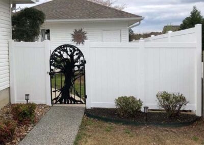 A white vinyl privacy fence with a black metal gate featuring a decorative tree design, set between landscaped garden beds with small shrubs, near a white house.