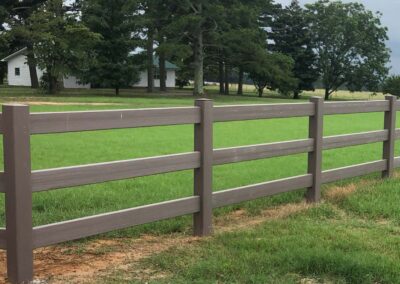A brown wooden fence runs alongside a green grassy field, with trees and a white house in the background under a cloudy sky.