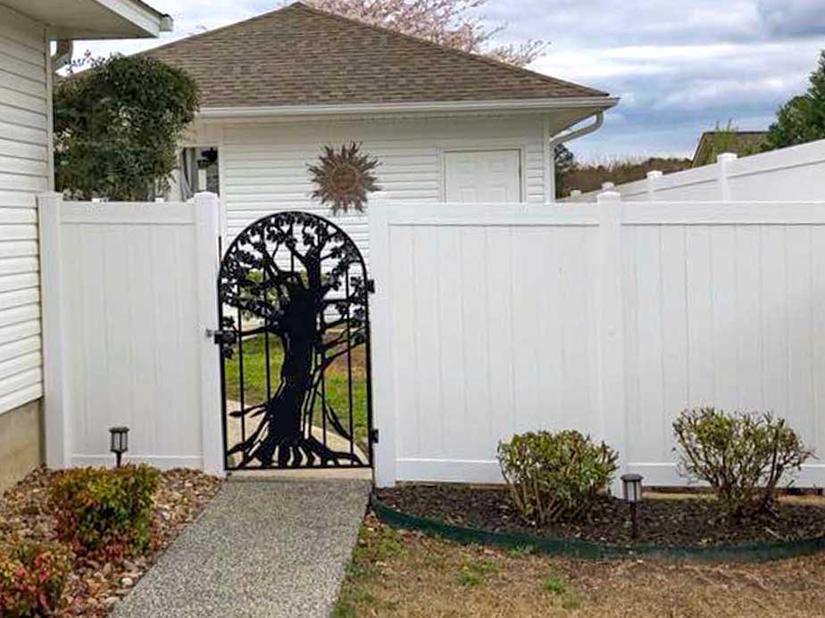 A white privacy fence with a decorative black metal gate featuring a tree design stands between a house and a garage. Shrubs line the fence, and a sun wall ornament hangs on the garage.