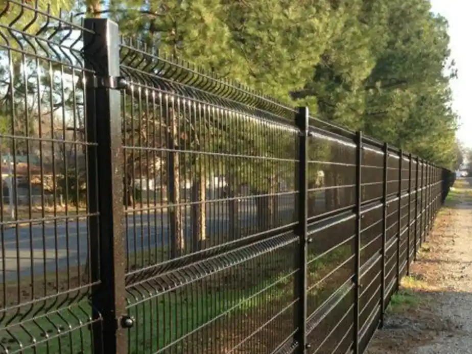 A tall black metal wire fence runs alongside a row of evergreen trees and a road, creating a barrier between the wooded area and the street.