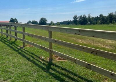 A wooden fence runs across a grassy field under a clear blue sky, casting long shadows on the ground. Trees and distant buildings are visible in the background.