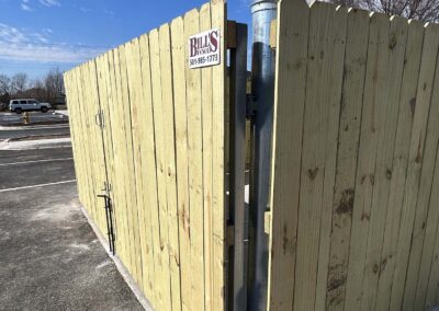 A tall wooden fence with a wide gate is slightly open, revealing a metal post inside. A sign reading Bills Fencing with a phone number is attached to the gate. The scene is in a parking lot under a clear blue sky.
