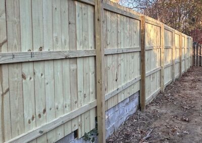 A newly built wooden privacy fence with vertical planks and horizontal supports stands on top of a short concrete block wall, with dry leaves scattered on the ground nearby. Trees are visible in the background.