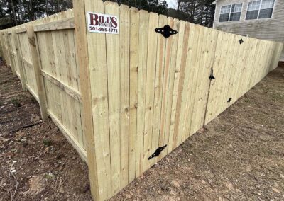 A newly built wooden privacy fence with black metal hinges is shown enclosing a yard. A sign reading “Bill’s Fences 501-985-1773” is attached to the fence. A house is visible in the background.