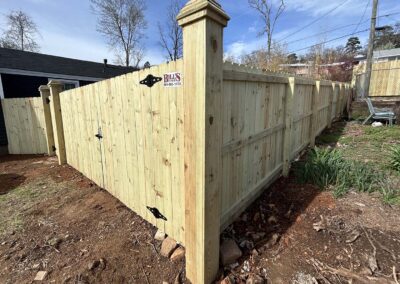 A newly constructed wooden privacy fence forms a corner in a yard. The fence is made of light-colored wood with black metal hinges and a small sign reading Bulls Fence Systems attached near a gate.