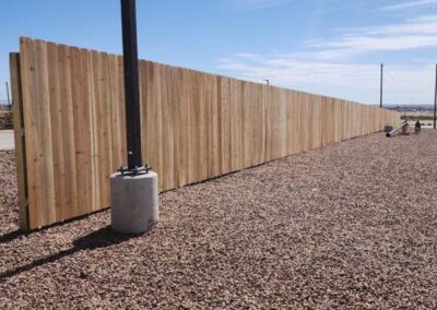 A long, wooden privacy fence stretches across a gravel-covered area on a sunny day, with a black utility pole set in concrete in the foreground.