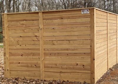 A large wooden privacy fence with horizontal planks and vertical support posts, set outdoors on a patch of dirt and surrounded by fallen leaves and bare trees. A small business sign is attached to the top corner of the fence.