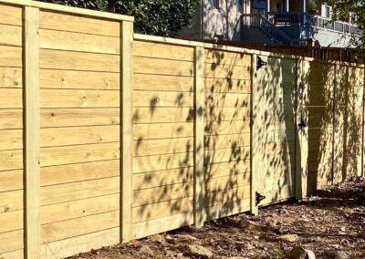 A newly constructed wooden fence with horizontal slats and a gate, casting shadows from nearby trees, stands on a patch of dirt and leaves with a house visible in the background.