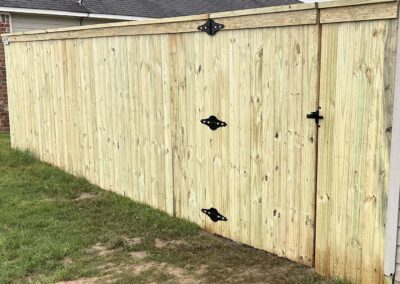 A wooden privacy fence with black metal hinges and a latch, standing on a grassy yard next to a house with brick and siding exterior. The fence appears new and well-constructed.
