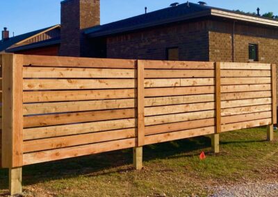 A horizontal wooden slat privacy fence stands in front of a modern brick house, with sunlight casting shadows on the fence and lawn.