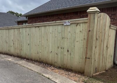 A tall wooden privacy fence with a small sign reading “BILLS” is shown in front of a brick house. The fence is along a paved alley, and the sky is cloudy.